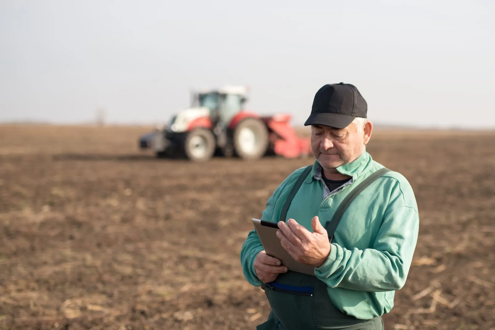 A farmer using a tablet beside a cultivated field and tractor